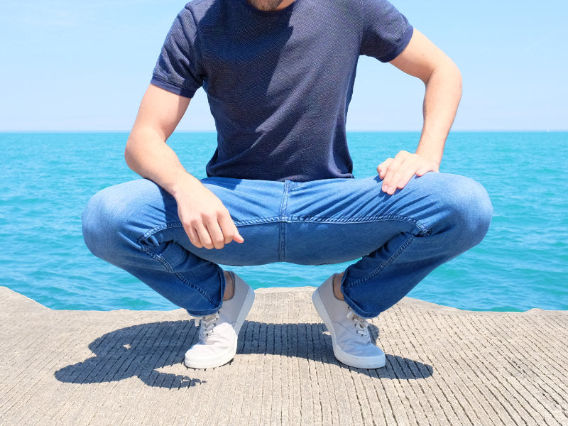A person squatting on a pier wearing light wash jeans and a navy shirt, with the ocean in the background.