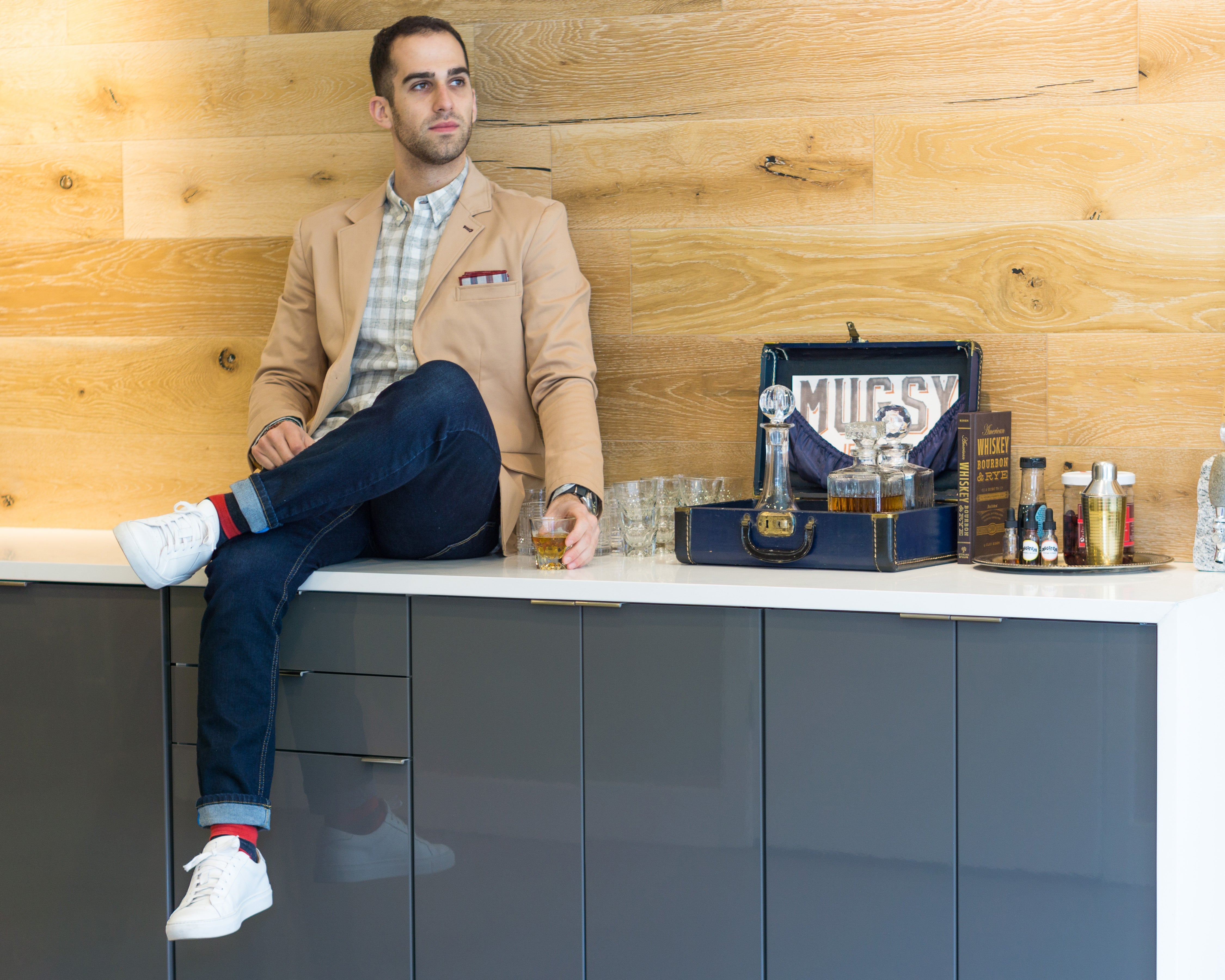 Man sitting on a counter wearing Mugsy stretch jeans, holding a drink, with a vintage suitcase and grooming products nearby.