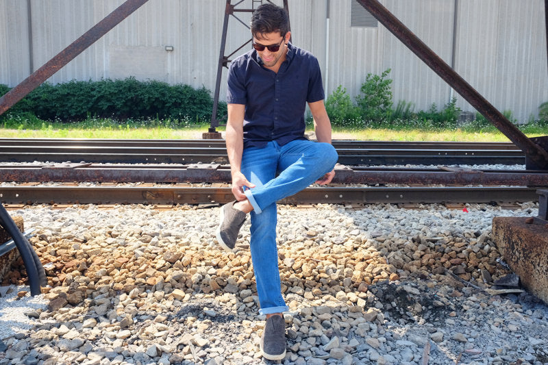 A man sitting on rocks, adjusting his jeans while wearing sunglasses.