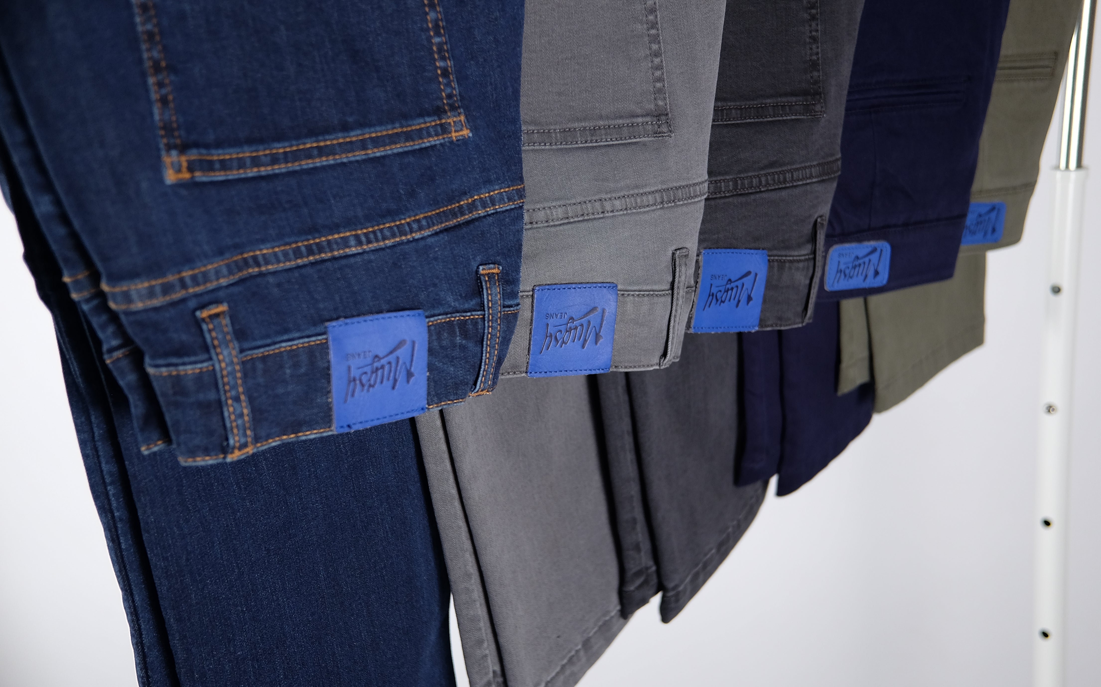 Mugsy jeans and chinos hanging, each with a blue patch labeled 'Mugsy'