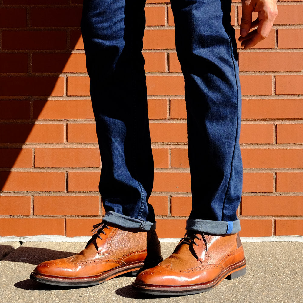 A person wearing brown shoes and cuffed blue jeans standing against a brick wall.