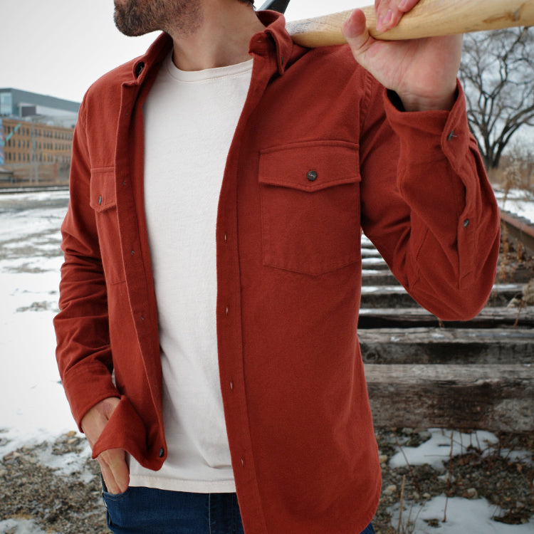 Man wearing a brick red flannel shirt, holding a baseball bat, standing outdoors.