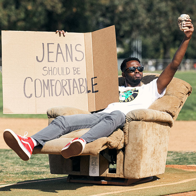 A person sitting in a recliner holding a sign that says 'JEANS SHOULD BE COMFORTABLE'.