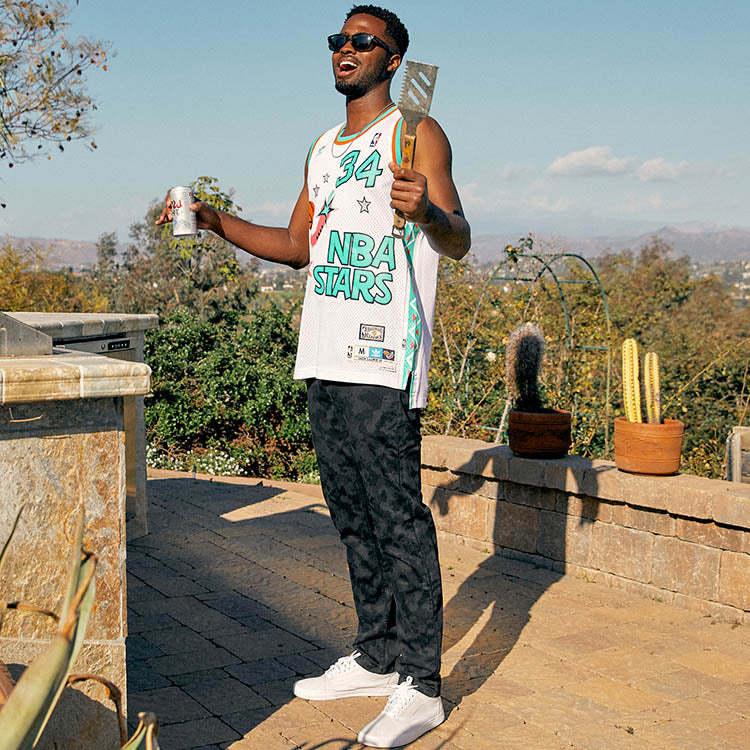 A person wearing an NBA jersey and sunglasses, grilling outdoors with a drink in hand.