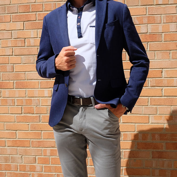 A person wearing a navy blazer over a white shirt and gray pants, standing against a brick wall.