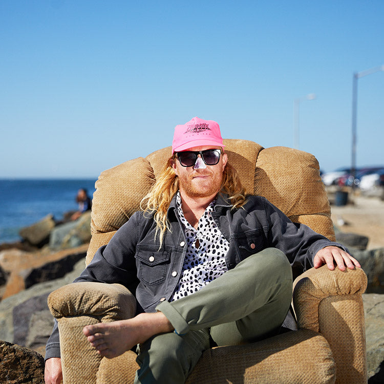A man wearing a pink hat and sunglasses sitting in a chair by the beach.