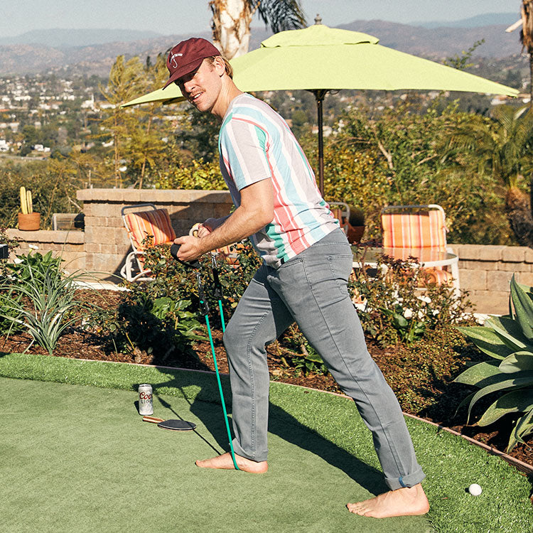 A man wearing a striped shirt and a maroon cap stands on a lawn, holding a golf club, with a can and phone nearby.