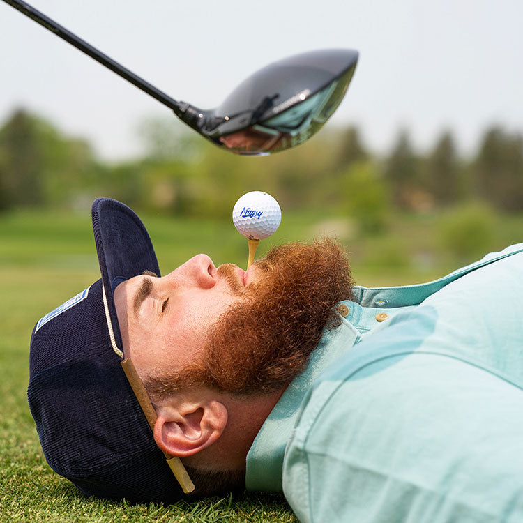 A man lying on the grass with a golf ball on his beard, while a golf club is poised above him.