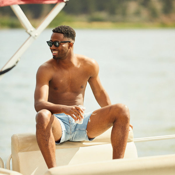 A man wearing denim shorts sitting on a boat, smiling and holding a drink.
