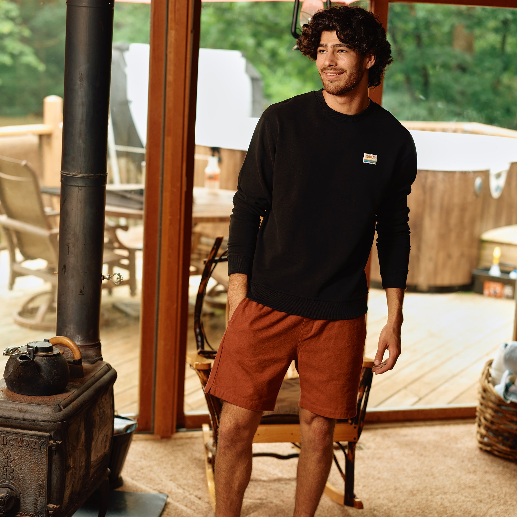 A man wearing a black sweatshirt and brown shorts, standing in a cozy indoor setting with wooden decor.