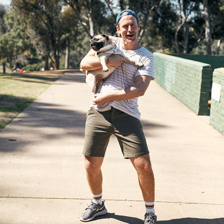 A man wearing a striped shirt and shorts holds a pug in a park setting.