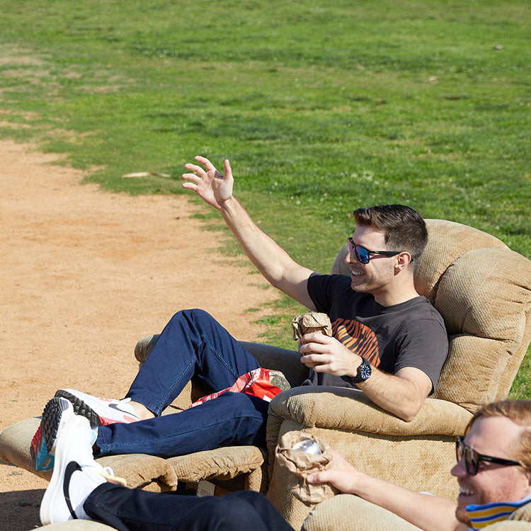 A man in sunglasses sitting in a chair, holding a drink and smiling.