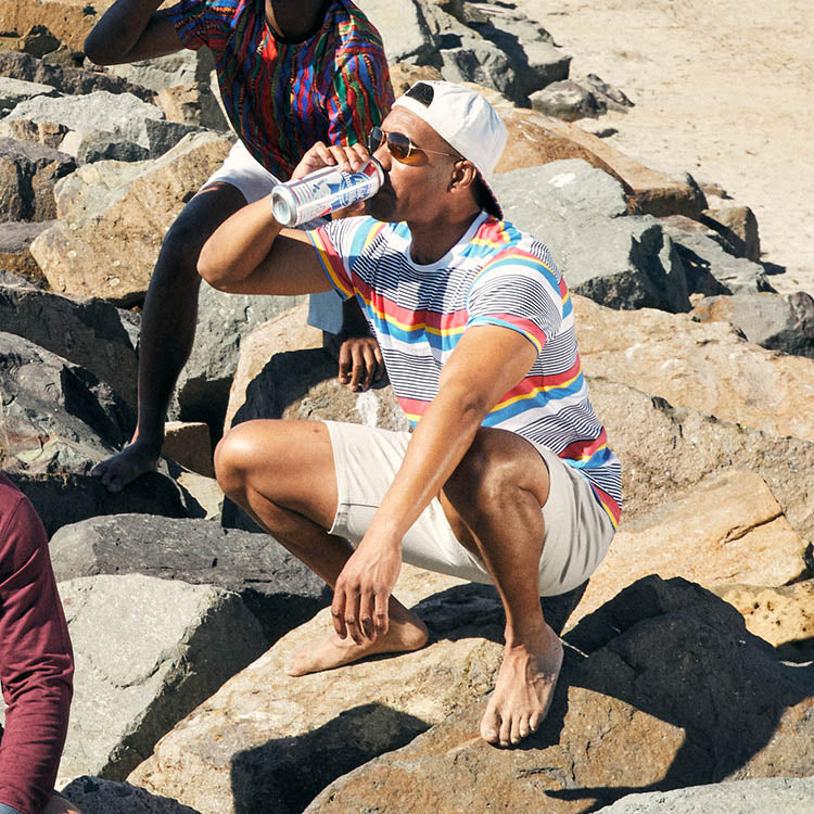A man in a colorful striped shirt squatting on rocks at the beach, drinking from a can.