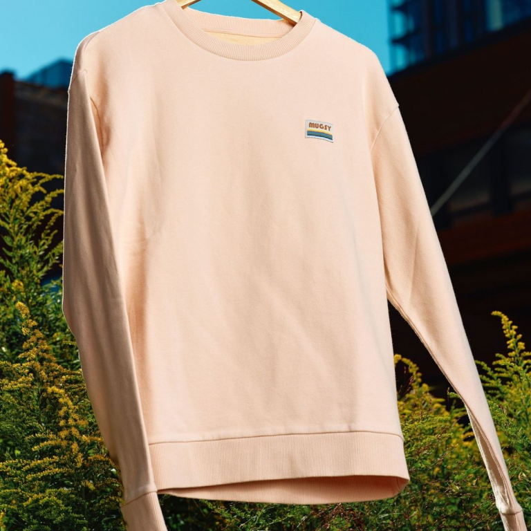Beige sweatshirt hanging on a wooden hanger outdoors with a patch reading 'MUGSY' on the chest.