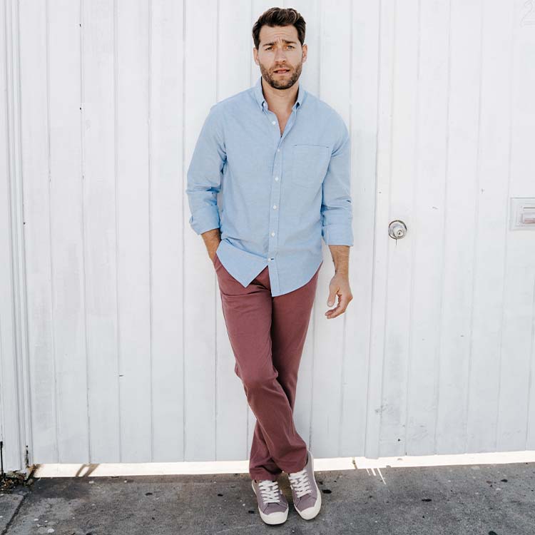 A man in a light blue shirt and maroon pants standing against a white wall.