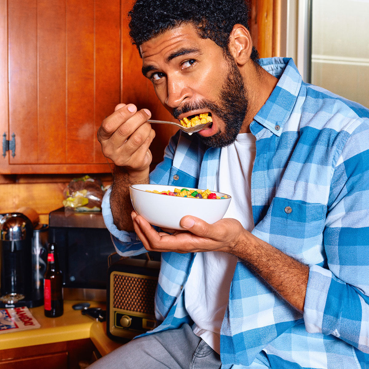 A man in a blue plaid shirt eating cereal from a bowl in a kitchen setting.