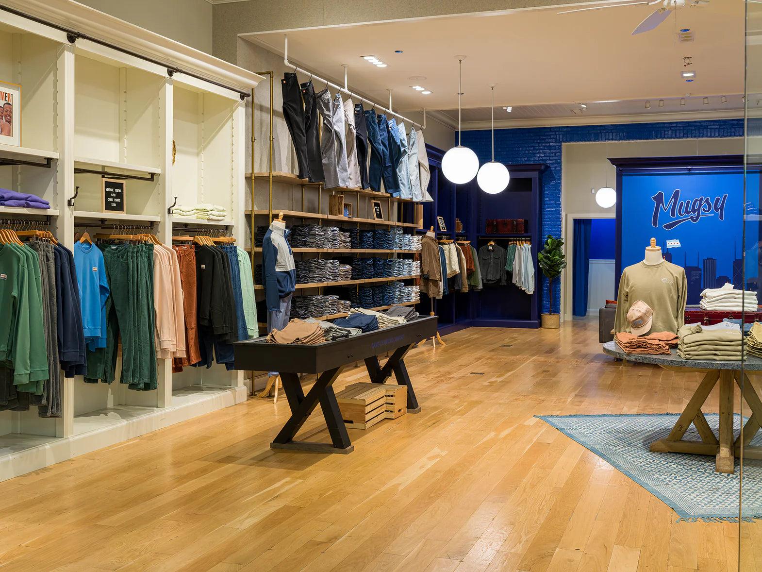 Clothing store interior with wooden floor, racks of pants and shirts, mannequins wearing casual outfits, and a blue wall with the store name 'Mugsy'.