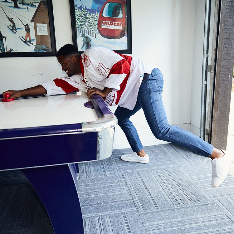 A person in a hockey jersey playing air hockey while wearing blue jeans and white shoes.