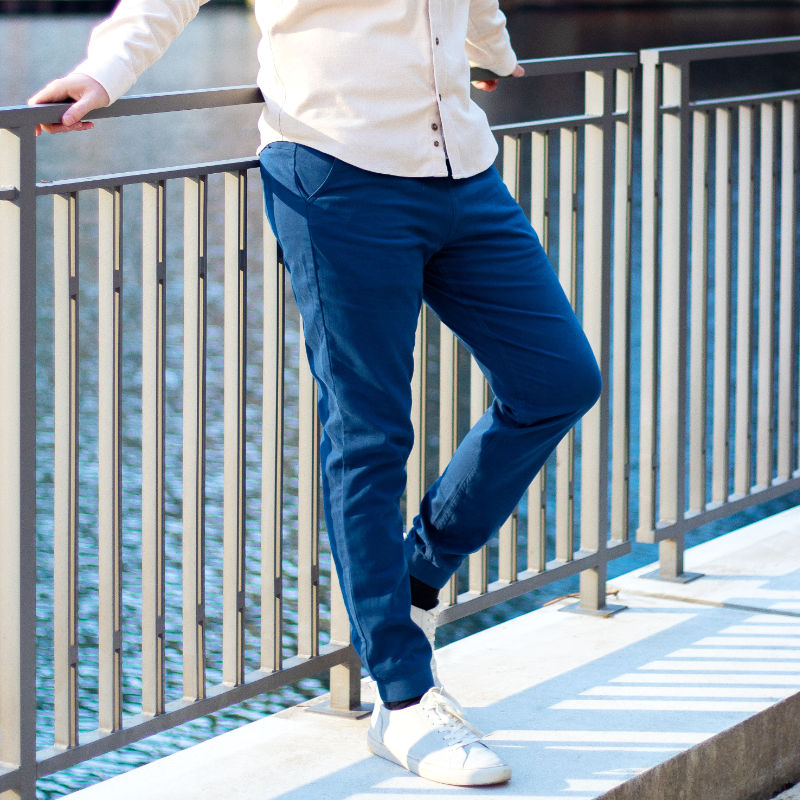 A person wearing navy joggers and white sneakers, standing by a railing near water.