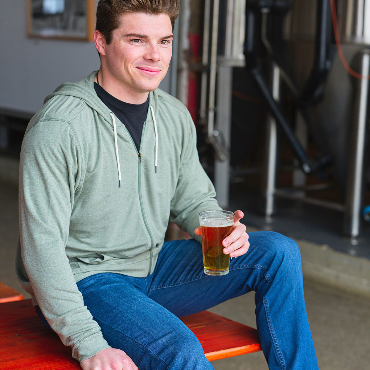 A man in a green hoodie sitting with a beer in a casual setting.