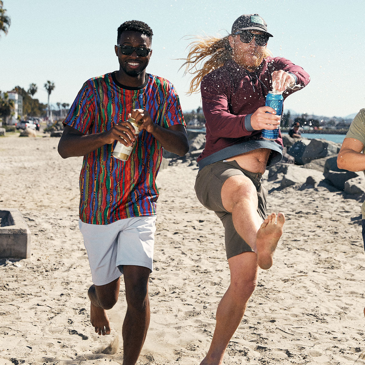 Two men on a sandy beach, one wearing a colorful shirt and white shorts smiling while holding a drink can, the other with long hair, sunglasses, and a cap kicking and spilling a blue drink can.