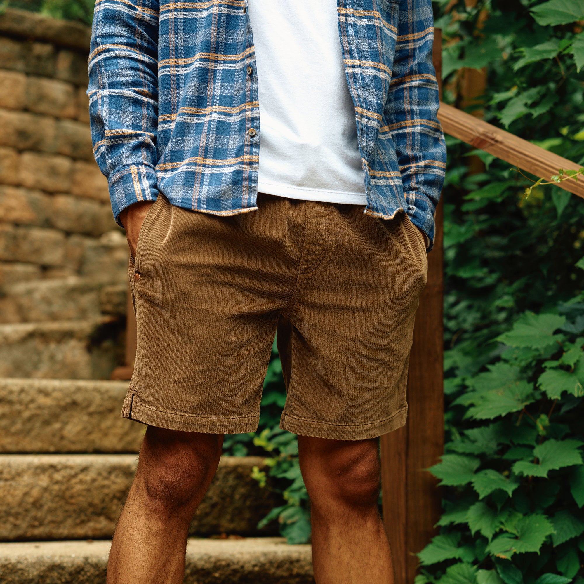 A person wearing brown corduroy shorts and a blue plaid flannel shirt, standing on steps surrounded by greenery.