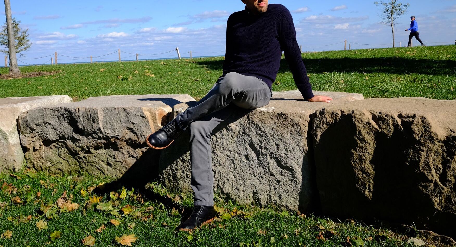 Man sitting on large rocks outdoors wearing a navy sweater, gray pants, and black boots on a sunny day with blue sky and green grass.