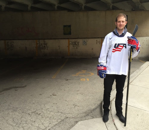 Man wearing a USA hockey jersey holding a hockey stick in a covered parking area