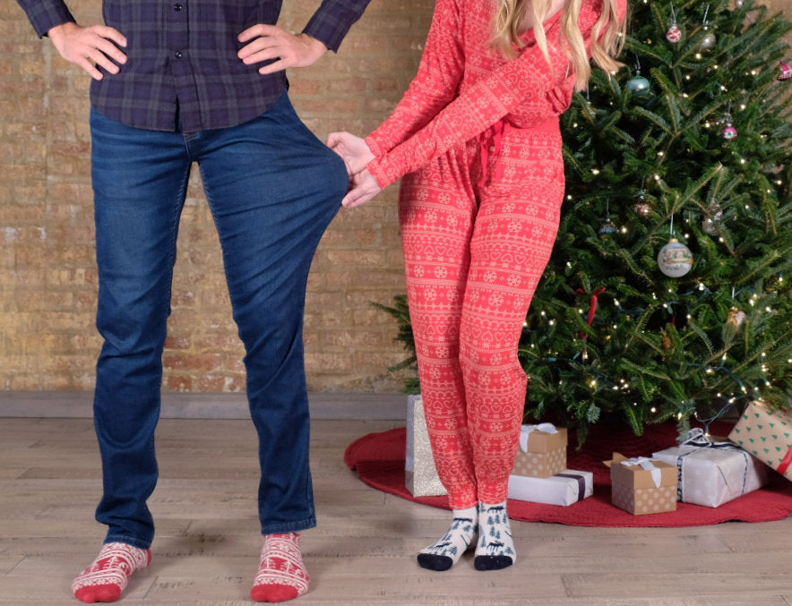 A man in plaid shirt and jeans, and a woman in red holiday pajamas, standing by a Christmas tree.