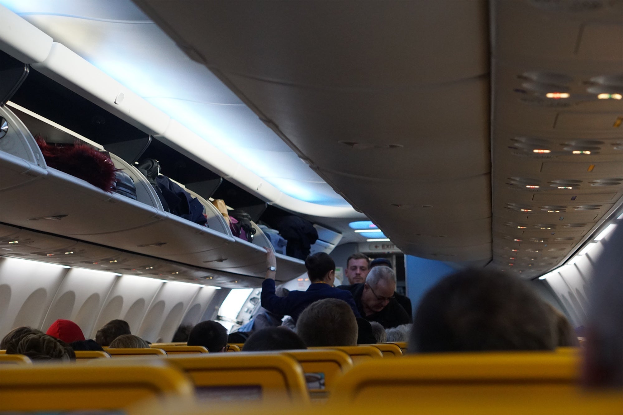 Interior of a budget airline cabin showing passengers seated and a flight attendant assisting with overhead bins.