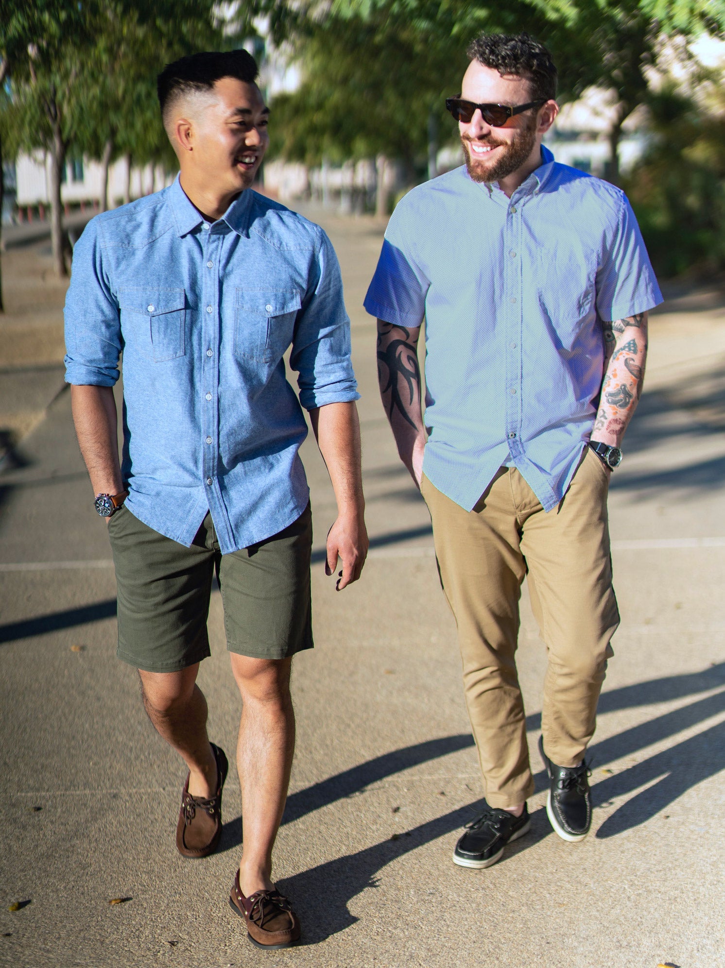 Two men walking, one in a denim shirt and olive shorts, the other in a light blue shirt and chinos.
