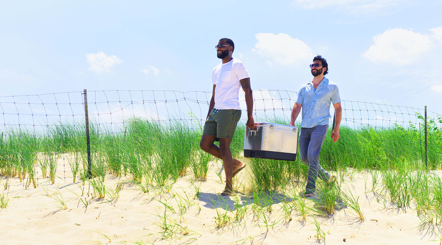 Two men walking on sandy beach grass carrying a metal cooler, wearing summer casual clothes: white t-shirt and green shorts, and light blue short-sleeve shirt with pants.