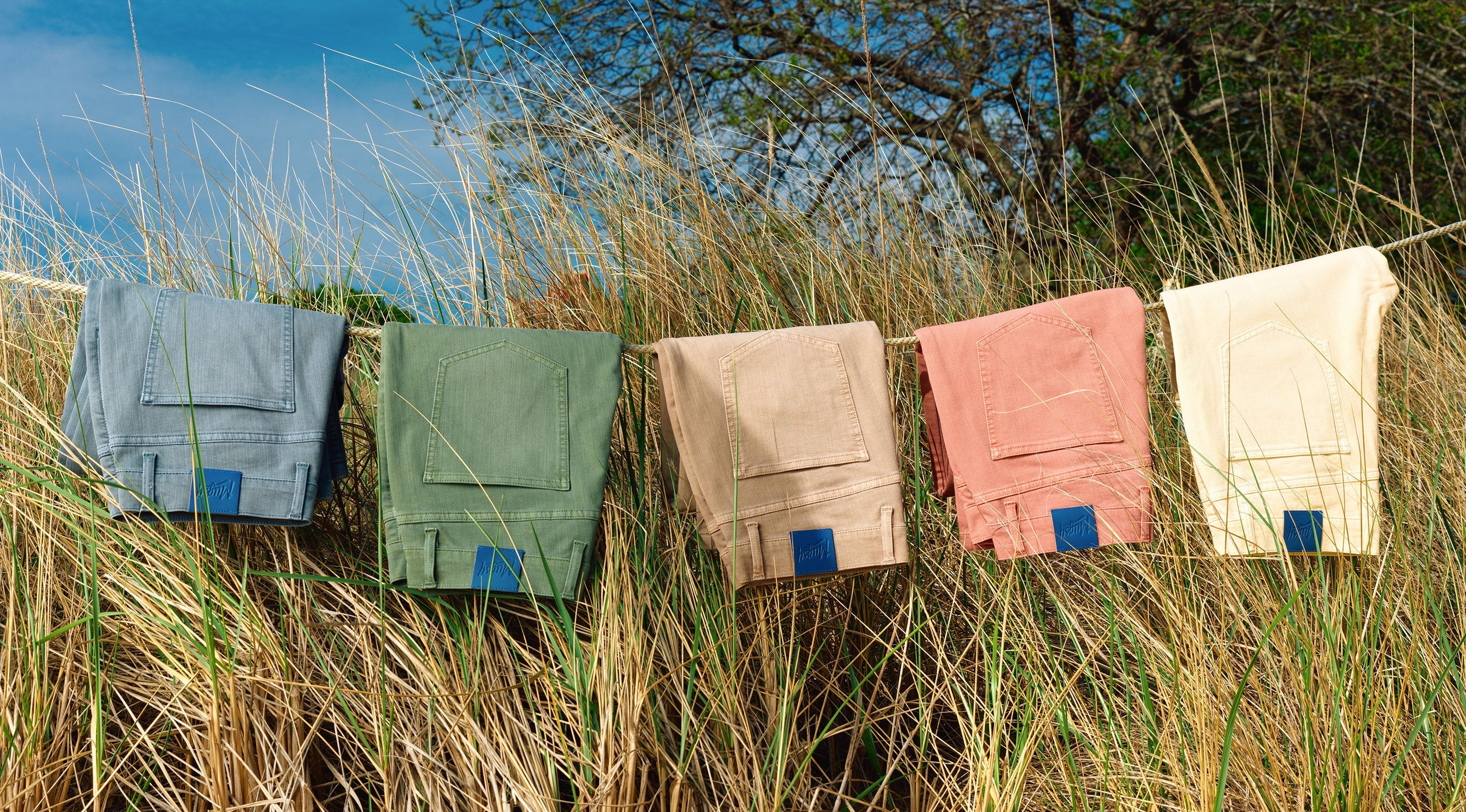 Five pairs of folded pants in blue, green, beige, coral, and cream hanging on a rope outdoors among tall grass under a clear blue sky.
