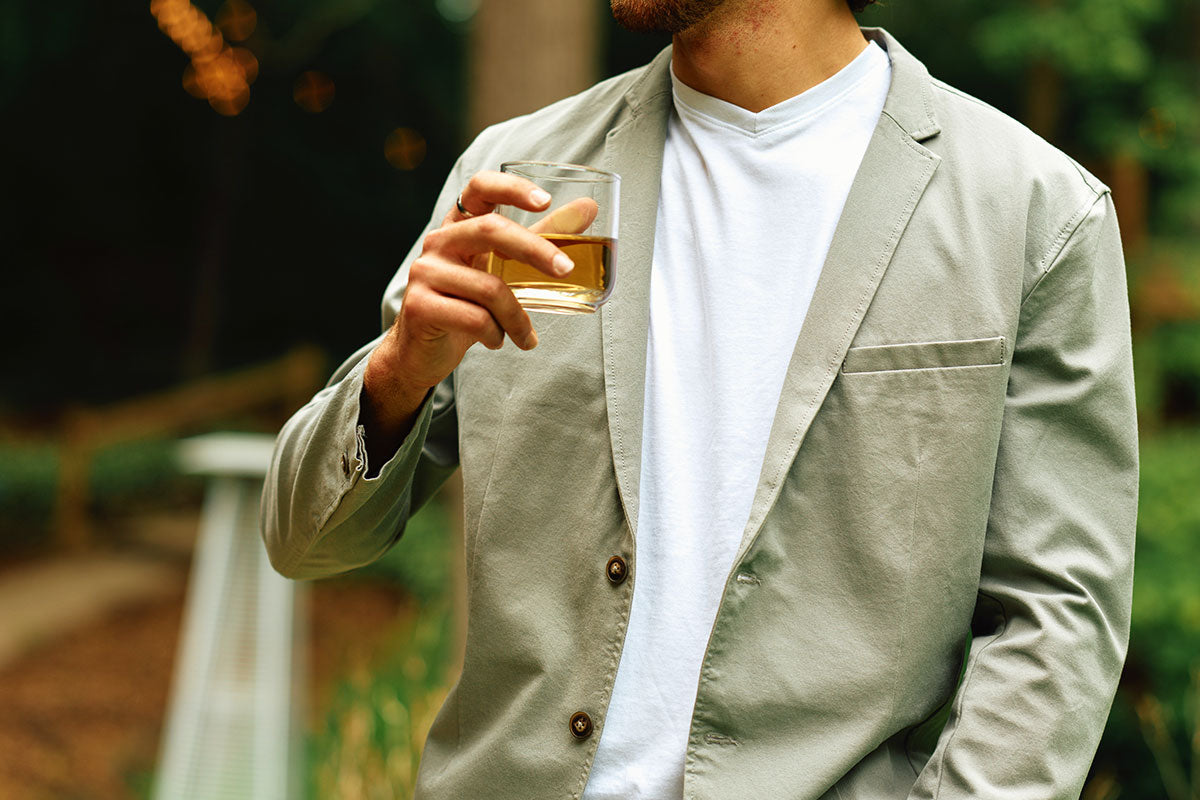 Man wearing a light beige blazer and white t-shirt holding a glass with amber liquid outdoors