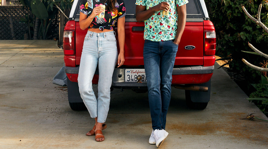 Two people standing and leaning against the back of a red Ford truck with a California license plate, wearing light blue and dark blue jeans, holding canned drinks, with colorful patterned tops and casual footwear.