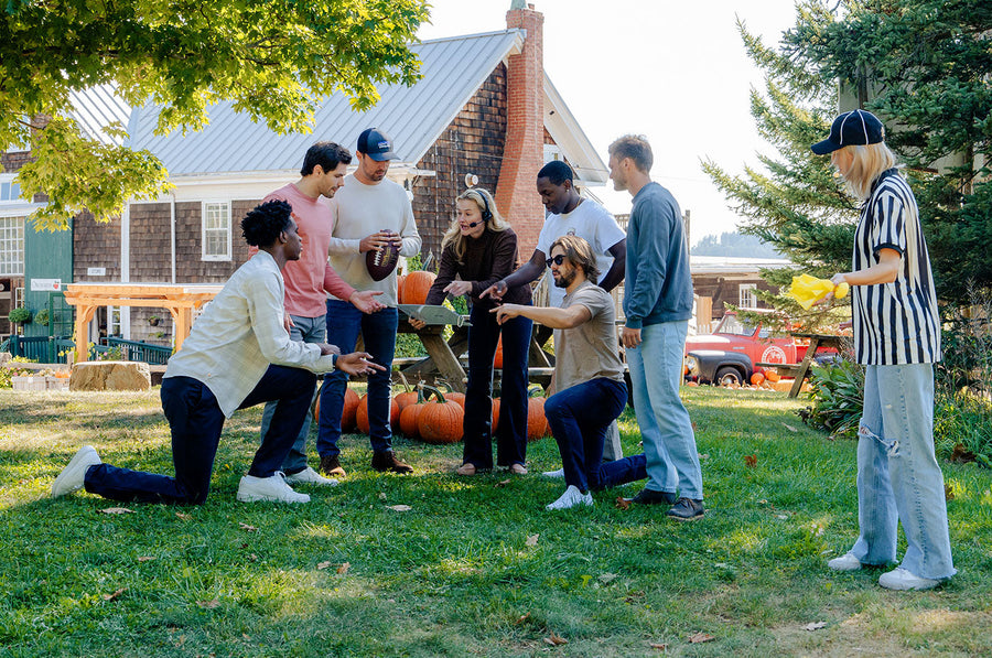 Seven people outdoors on grass near pumpkins and a rustic building, engaged in a casual football tailgate game; one person wears a referee shirt and holds yellow flags, others hold a football and point at a tablet device.