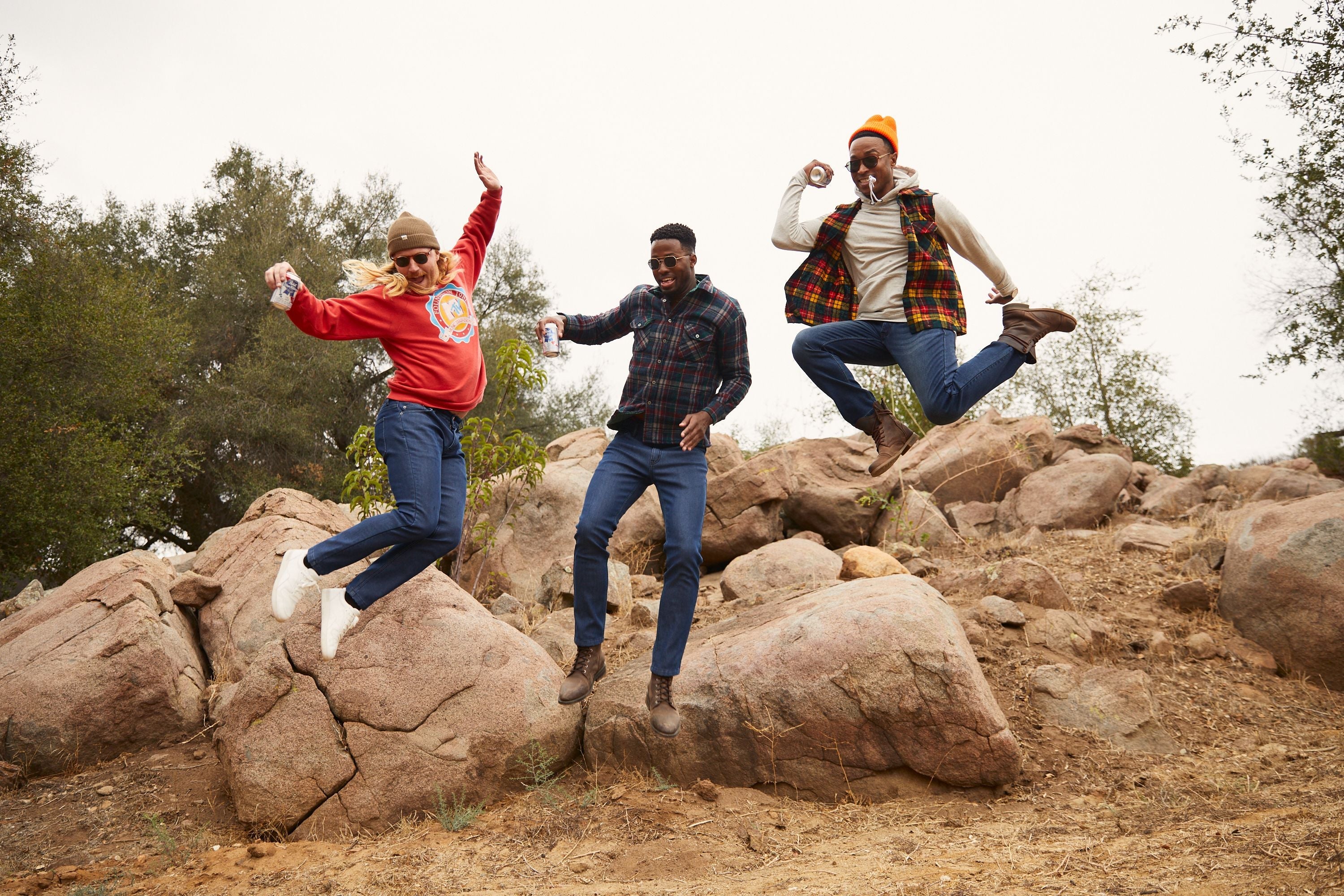 Three men jumping on a rocky hillside wearing jeans, flannels, and fall layers