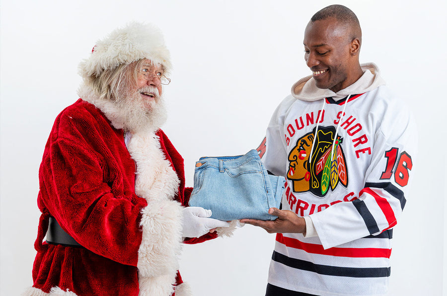 Santa Claus handing folded light-wash jeans to a smiling man wearing a hockey jersey against a white background