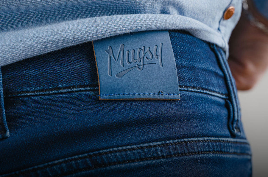 Rear close-up of Mugsy jeans showcasing embossed leather logo patch and dark indigo denim texture