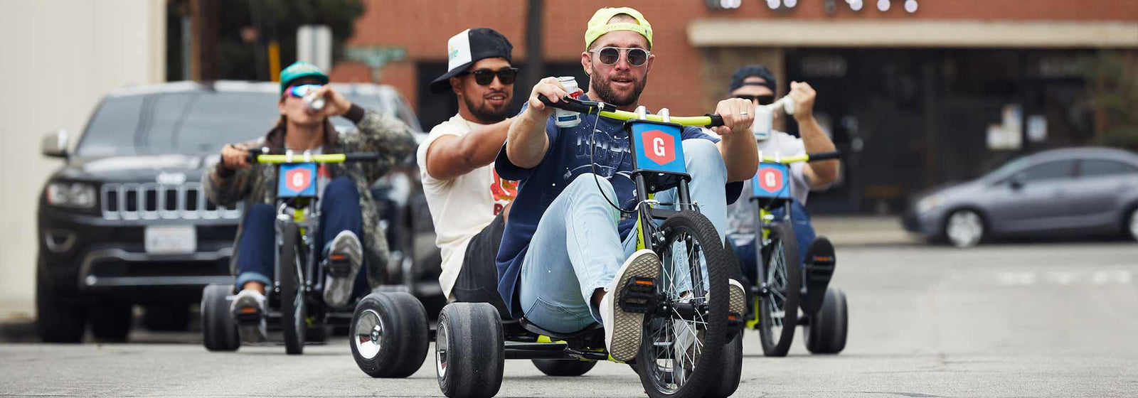 Three men riding tricycles on a street, each tricycle displaying a blue plate with a red shield and white letter 'G'. The men wear casual clothes and sunglasses, and one man in front wears a yellow cap backwards.