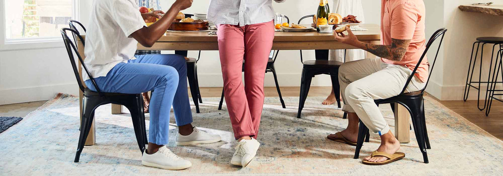 Three people seated at a dining table showing lower bodies: colorful pants, white sneakers and sandals on a patterned rug.