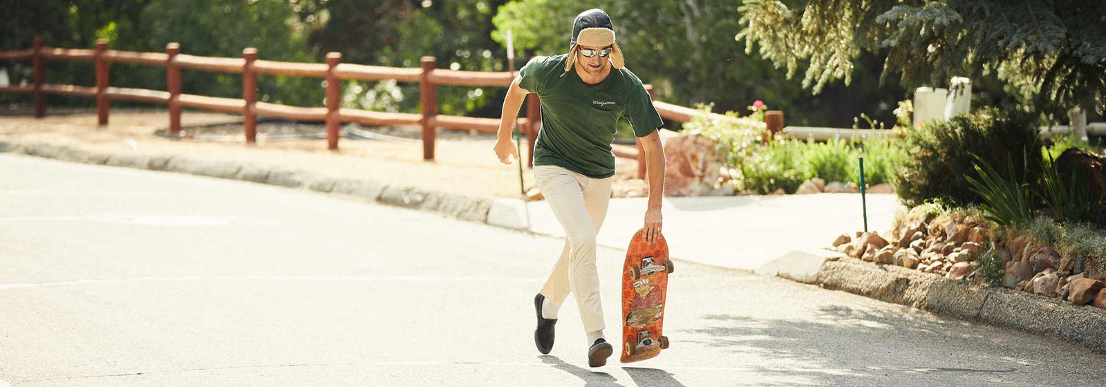 Man wearing a green shirt and beige pants running on a sunny street while holding an orange skateboard with stickers