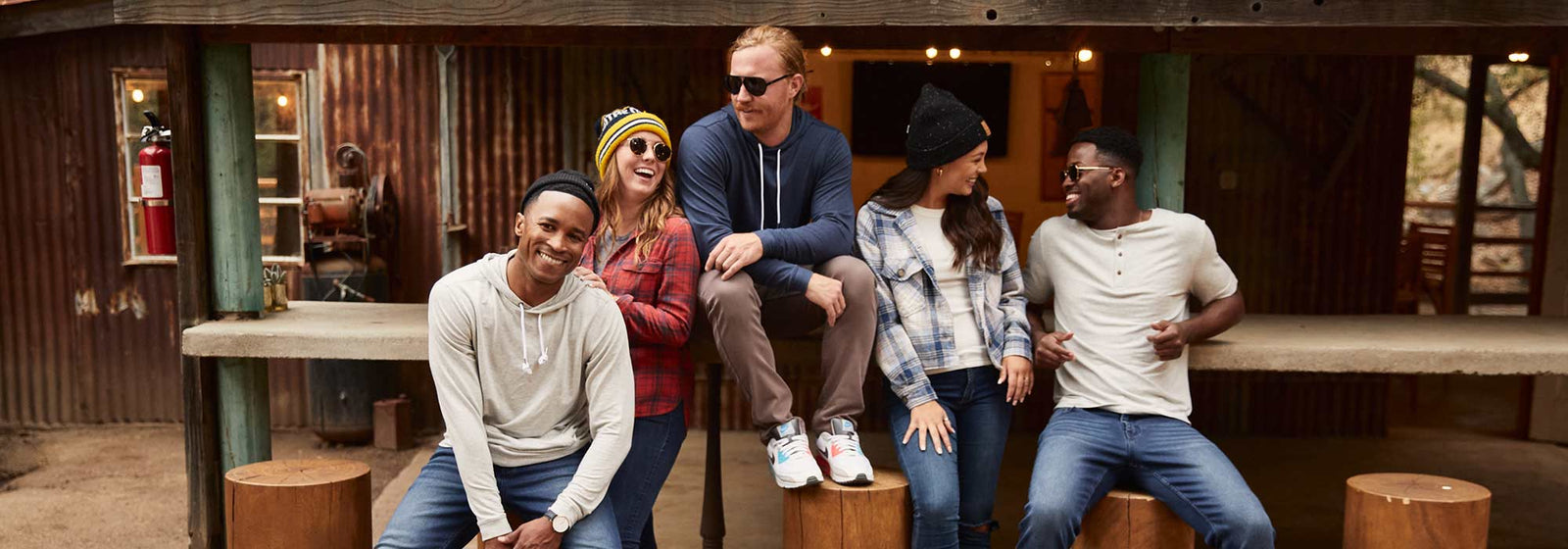 Five people wearing casual tops and jeans, sitting and standing on wooden stools in a rustic outdoor setting, smiling and interacting.