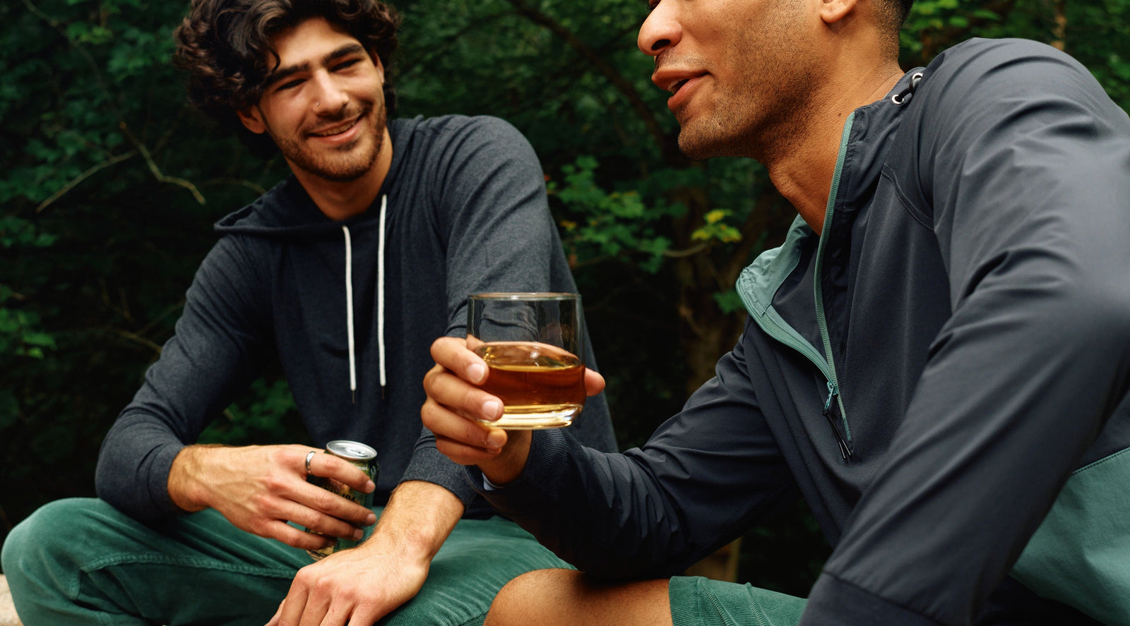 Two men outdoors wearing Cashtech™ hoodies, one holding a glass with amber liquid and the other holding a can, smiling and conversing.