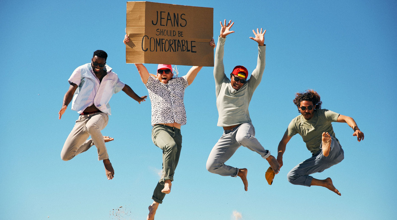 Four men jumping in the air against a blue sky, one holding a cardboard sign that reads 'JEANS SHOULD BE COMFORTABLE'