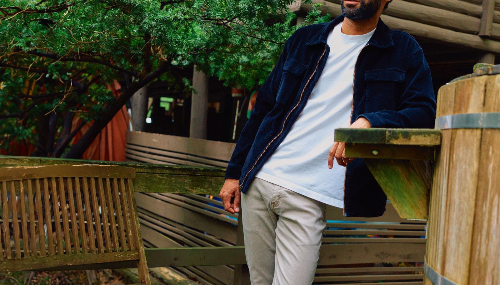 Man wearing a navy corduroy jacket and white t-shirt leaning on a wooden railing outdoors with greenery in the background