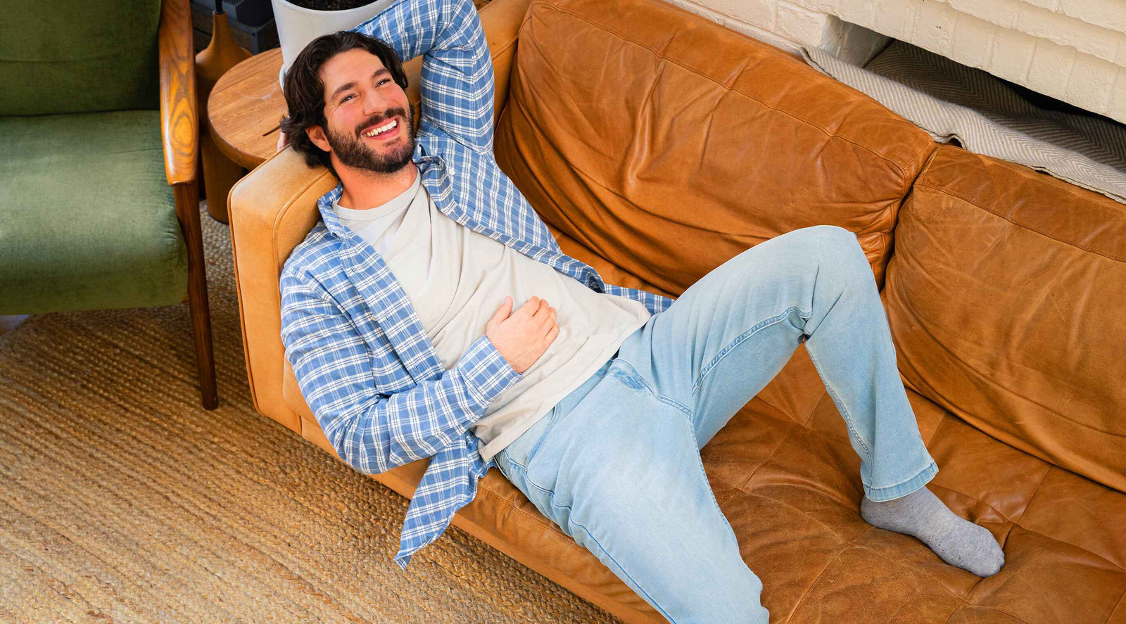 Man reclining on a brown leather couch wearing a blue plaid flannel shirt, light jeans, and gray socks in a cozy living room setting.
