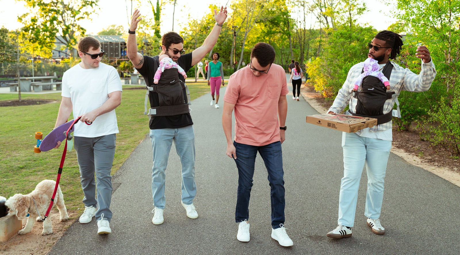 Four men walking on a paved path outdoors, two carrying babies in front carriers, one holding a skateboard, and one holding a pizza box, with trees and people in the background.