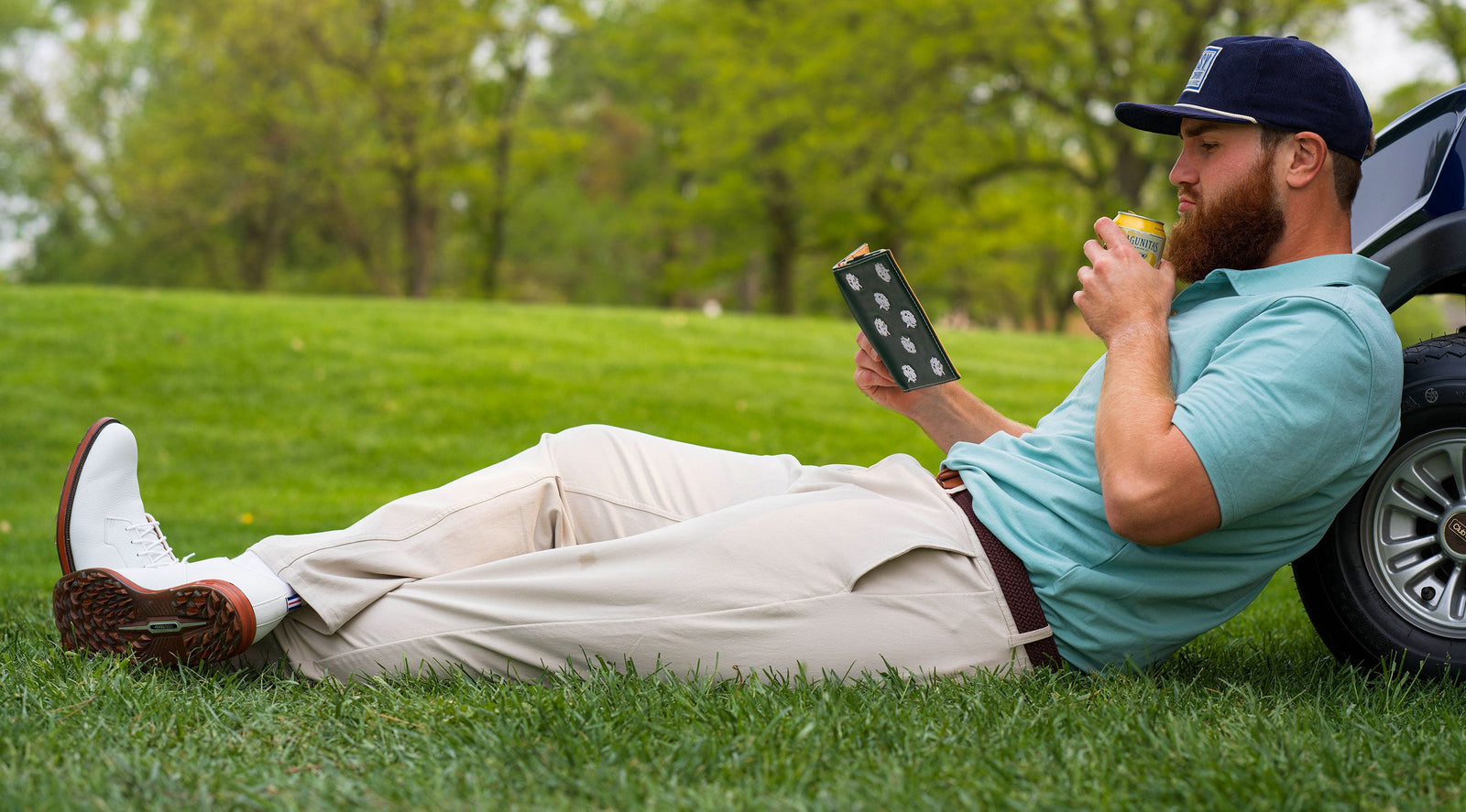 Man reclining on grass beside a golf cart, wearing beige golf pants, white golf shoes, a teal polo shirt, and a navy cap, reading a book and drinking from a can.