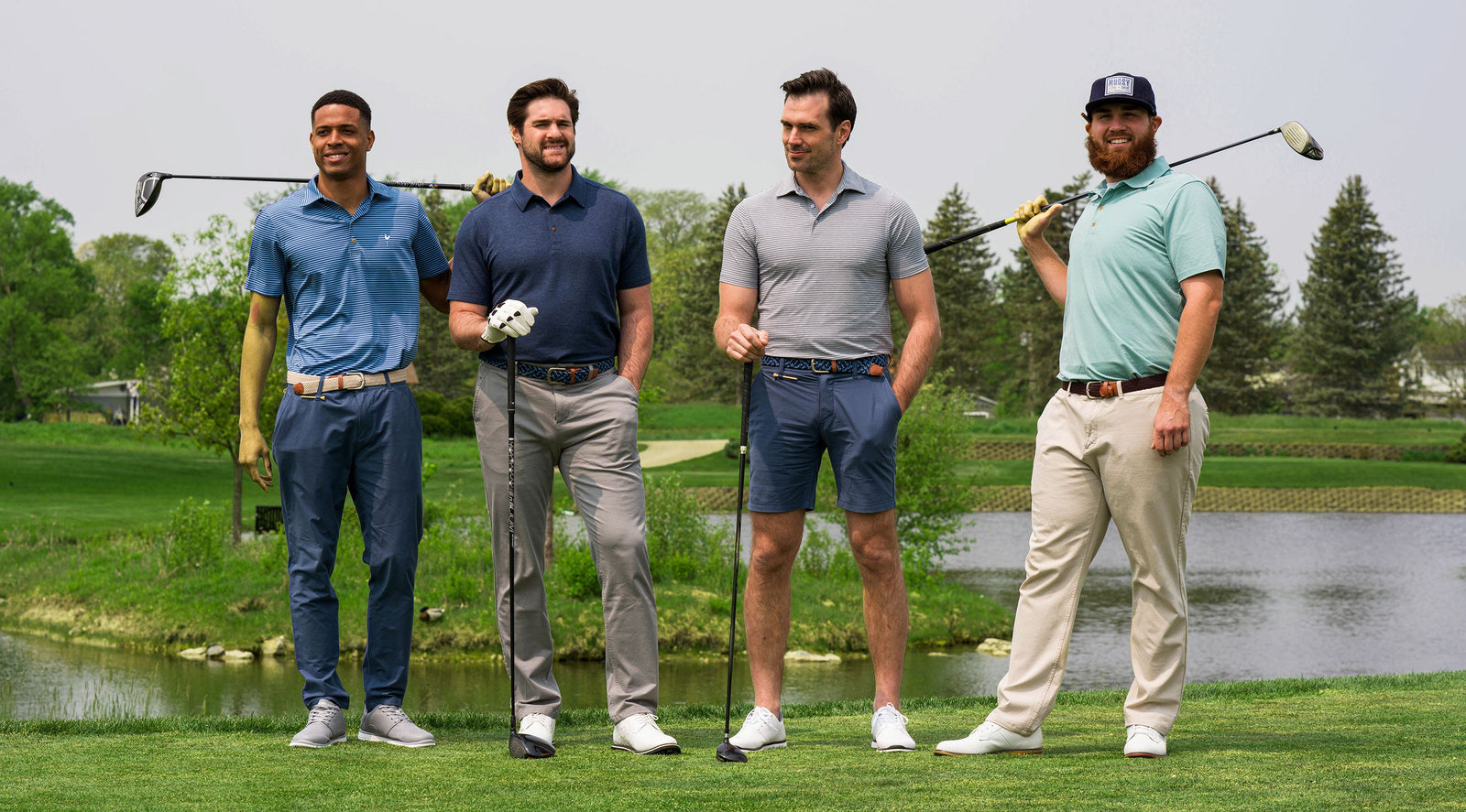 Four men standing on a golf course holding clubs, wearing Mugsy performance golf shorts and pants in navy, gray, and khaki